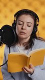 Vertical video Narrator wearing headset reading aloud from book into mic against yellow background. Upbeat professional voice actor recording audiobook, creating engaging media content for listeners