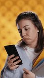 Vertical video Upset woman holding stack of book, preparing for school exam using phone, studio background. Student with pile of textbooks pouting while researching for university homework using