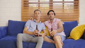 Caucasian attractive young gay couple watch football match on television. Attractive two male LGBTQ friend having fun cheering for their team while sitting on sofa watch soccer on TV together in house