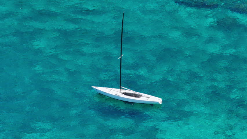 A small sailboat bobs gently in the vast expanse of the ocean, with no land in sight. The sailboat appears to be navigating through the open waters, surrounded by nothing but blue waves and sky.