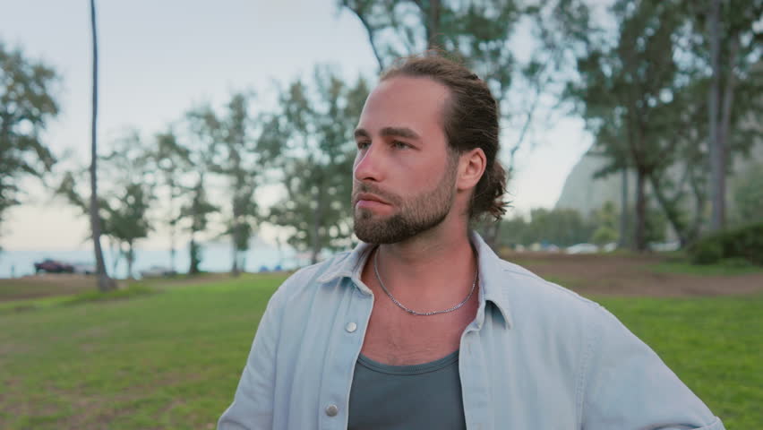 A solitary man enjoys a moment of reflection, holding a cigaret in a park setting, surrounded by the beauty of serene nature and the quiet of the evening. Slow motion. 