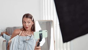 Young social medial content creator woman make fashion video. Blogger smiles to camera using selfie stick and light ring while making persuasive online clothing sell vlog to audience or follower. Adit