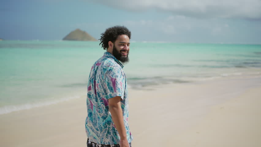 With a joyous smile, a man in a colorful tropical shirt and shorts walks along a sun-drenched beach, the turquoise ocean and distant islands creating an idyllic scene. Hawaii, Oahu, Slow motion.  Royalty-Free Stock Footage #3491025477