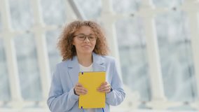 Playfull and Happy young businesswoman with curly hair and glasses is holding a yellow note book, her cheerful smile complementing her casual professional attire in a contemporary glass structure 