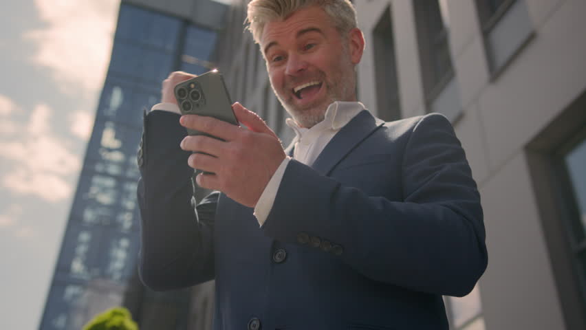 Amazed happy joyful success old senior Caucasian man mature middle-aged male employer business entrepreneur businessman in city downtown mobile phone win achieve prize victory good news in sunshine