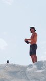 Fitness trainer drinks water from a bottle. Male athlete on the mountain against the sky. Outdoor sports training. Vertical video.