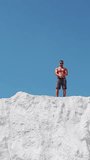 A strong man with glasses demonstrates his muscular body. Bodybuilding on the street on a white mountain against the blue sky. Vertical video.