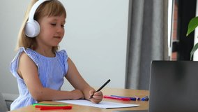 Little schoolgirl nods head and writes on paper during video lesson at home. Diligent junior student does homework at laptop. E learning services