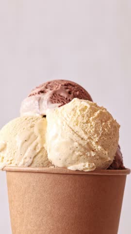 Chocolate sauce flowing on Chocolate and vanilla ice cream scoops in paper take away cup rotating over light background. Sweet dessert, rotation closeup. 4K 