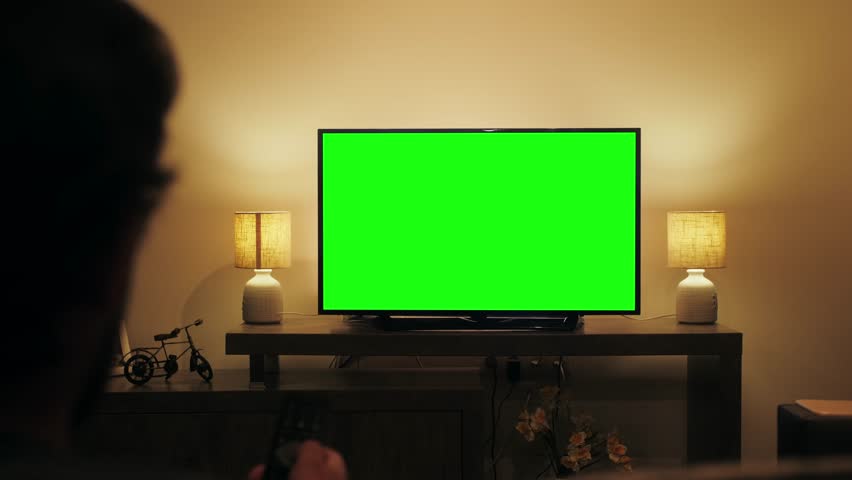 Person watching television with chroma green screen. Unrecognizable female person switching channels on tv with remote controller, sitting on sofa in living room.