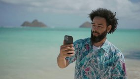 Young man with a joyful expression and curly hair taking video selfie on a tropical beach, clad in a patterned shirt with the serene turquoise ocean and clear sky in the background. Slow motion. 