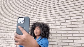A lively slow-motion video clip features a youthful African American woman with curly hair capturing selfies against a textured brick wall background. She playfully adjusts her hair, poses, and blows