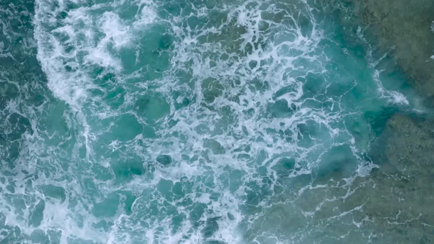 Top Down Slow Motion Drone View of turquoise clear water and crashing waves over shallow coral reef in Uluwatu Bali Indonesia. Clear water and abstract wave action.