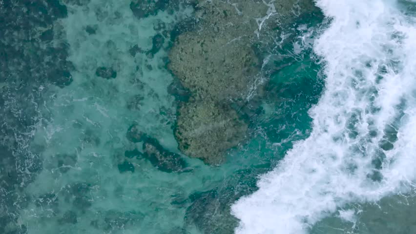 Top Down Slow Motion Drone View of turquoise clear water and crashing waves over shallow coral reef in Uluwatu Bali Indonesia. Clear water and abstract wave action.