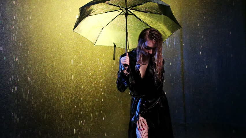 Female poses slightly leaning forward under pouring rain covering up with umbrella. Woman stands under rainfall in light of street lamp