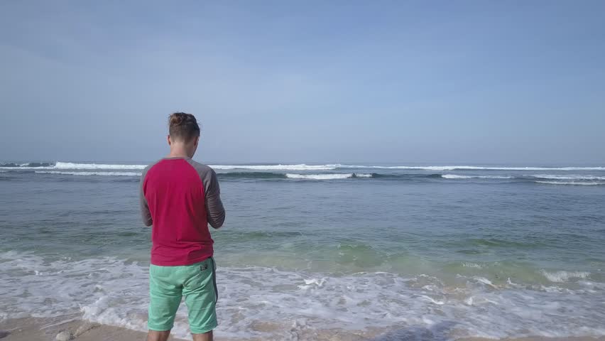 Young man operating quadcopter drone using remote control on seashore. Filming surfers on waves from tropical sand beach. Summer vacation and travel.
