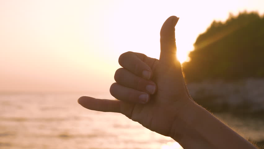 LENS FLARE, CLOSE UP, SILHOUETTE: Shaka hand gesture against golden setting sun expressing good vibes and aloha spirit. A positive hand gesture lit by a beautiful sunset symbolizes holidays on a beach