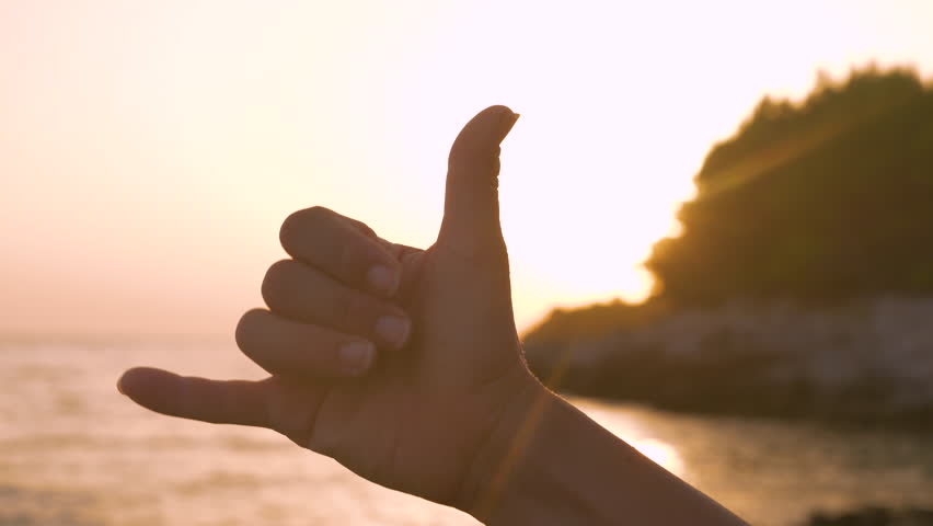 LENS FLARE, CLOSE UP, SILHOUETTE: Morning sunrise on beach and shaka hand gesture expressing good vibes and aloha spirit after fun surf session. Positivity and happiness on summer holidays at seaside.