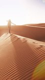 Vertical video. A person, possibly a girl, is standing on the peak of a sand dune. The vast desert landscape stretches out behind her, showcasing the rugged beauty of the dunes under the bright sun.