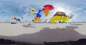 The video provides a 360-degree view of a beach filled with people enjoying a kite festival. The sky is adorned with an array of vibrant kites in various shapes and sizes, including a red bird, a