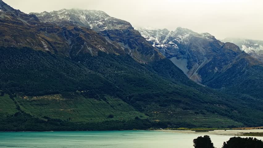 Misty layer of clouds across snow capped mountains overlook calm waters in New Zealand