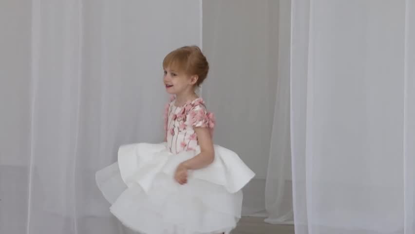 A young girl holds a bouquet of flowers in her hand. She is wearing a white dress and her hair is in a bun. The action takes place in a room with white walls and a white curtain.