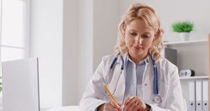 Concentrated professional young female medical doctor in white uniform with stethoscope working at the desk in office on a laptop making notes in notepad. Physician woman working in clinic. 4k video.