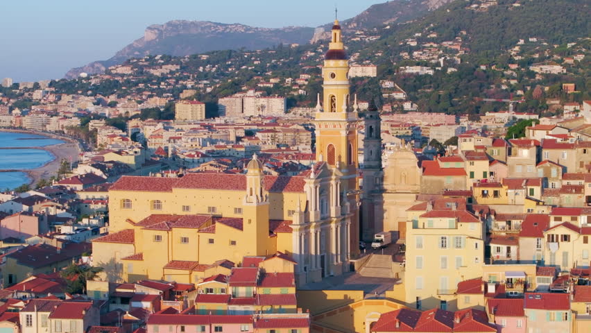 Aerial: A Riviera Town Awakens, With The Rising Sun Casting A Soft Light On The Ornate Church And Multicolored Facades, Against The Calm Sea. - Menton, France