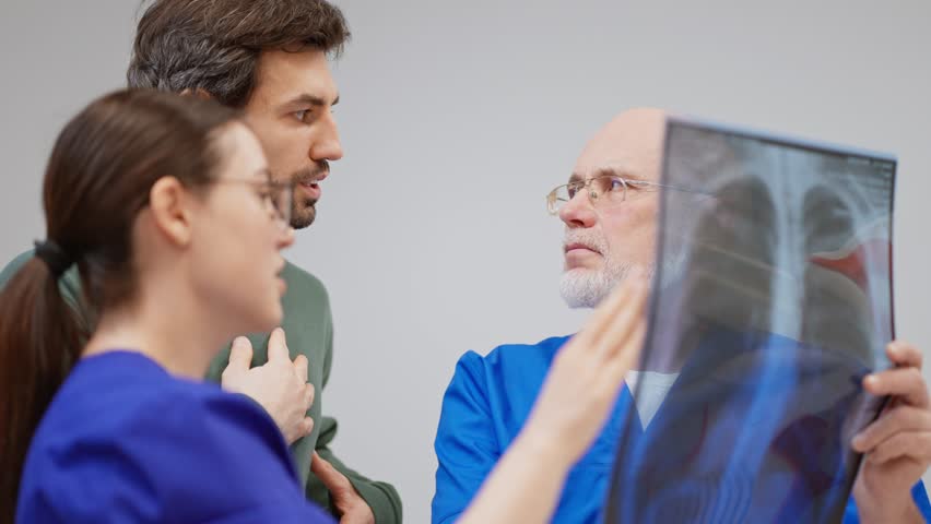 A brunette guy with stubble in a green jacket talks to an experienced doctor with a beard in a blue uniform and a brunette girl doctor about his chest fluorography in a modern clinic