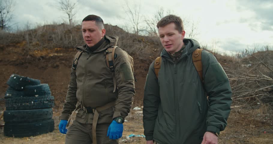 Two confident military men in green uniforms and with backpacks on their backs walk along a combat training ground with a barricade of car tires and communicate in the steppe Royalty-Free Stock Footage #3492064105