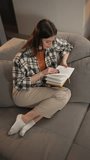 Vertical video from above of a confident and calm brunette girl in a checkered shirt and an orange T-shirt sitting on a gray sofa and reading a book at home in a modern apartment
