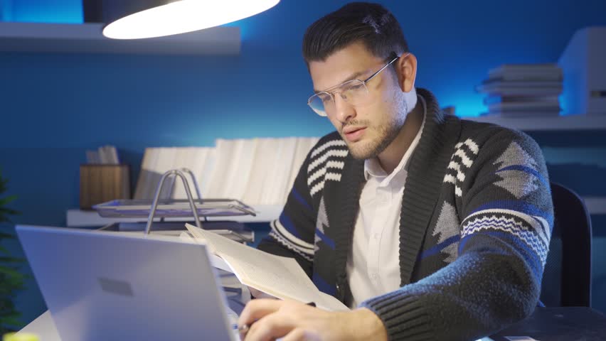 Young historian man reading a book at night in his study, typing his research on the laptop.