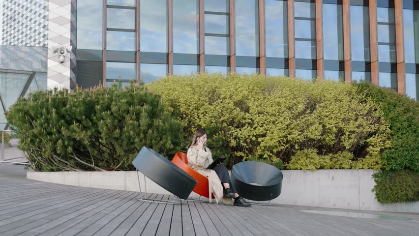 Young businesswoman in a white shirt and coat with a folder in her hand talking on the phone in front of office buildings