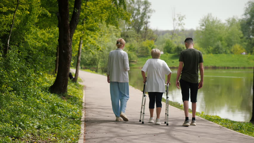 An elderly woman walks in the park with the help of a walker, her daughter and grandson accompany her nearby. Back view