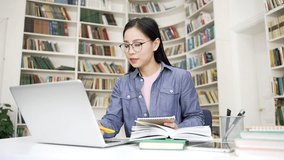 Asian female student listens to online video course using a laptop computer while sitting in campus library space. A girl at a remote learning seminar or lecture writes in a notebook in the classroom