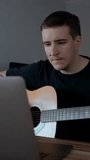 Man watches online lessons on laptop learning to play guitar. Male person with musical instrument sits at wooden table near gadget slow motion Vertical footage.