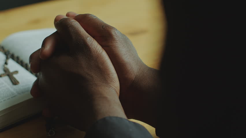 Clasped hands of unrecognizable Black clergyman leaning on wooden table with opened Bible book while praying. Close-up view