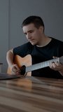 Man plays guitar watching educational videos on laptop. Beginner musician learns to use musical instrument sitting at wooden table slow motion Vertical footage.