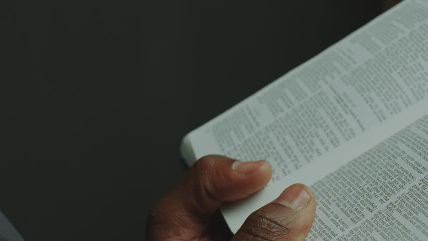 Black pastor in cassock holding opened Bible and reading a prayer in catholic church. Close-up view, tilt-up shot