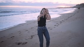 Slow motion of cute silly young woman run around beach on sunset making selfies on smartphone or talking through video chat conversation app