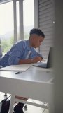Vertical video of teenage boy sitting at desk in bedroom at home doing homework on laptop - shot in slow motion