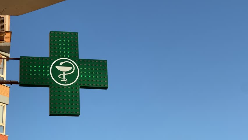 Green LED-Illuminated Pharmacy Cross Sign Mounted on Apartment Building Against the Blue Sky