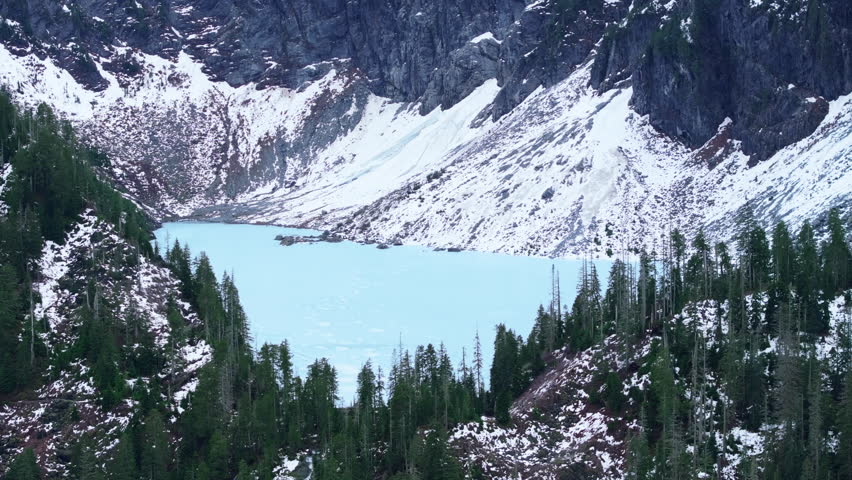 An icy blue lake lies frozen at the base of a snow-draped mountain, surrounded by dense evergreen forests in the heart of Washingtons rugged terrain. 4K footage.  Royalty-Free Stock Footage #3492399191
