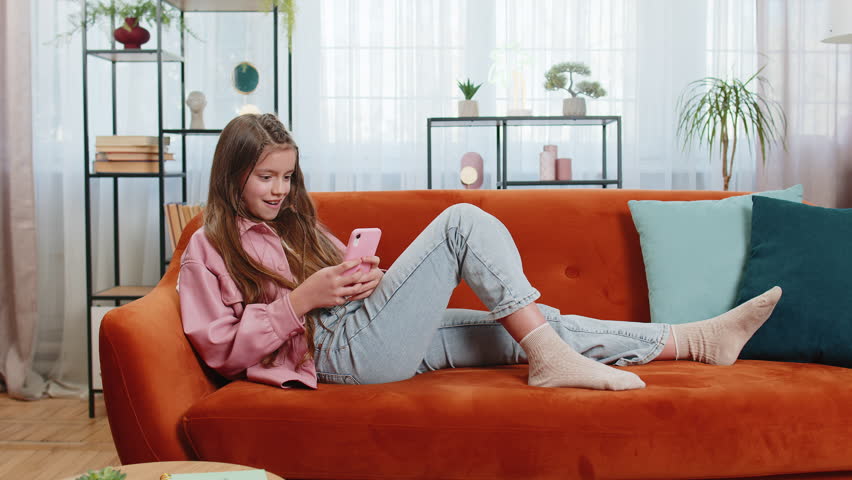 Young girl texting share messages content on smartphone social media applications online watching relax movie. Female Caucasian teenager kid child uses mobile phone sits on sofa in living room at home