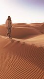 Vertical video. A man walks across a vast sandy desert field, with dunes visible in the background. The barren landscape stretches out endlessly under the scorching sun.