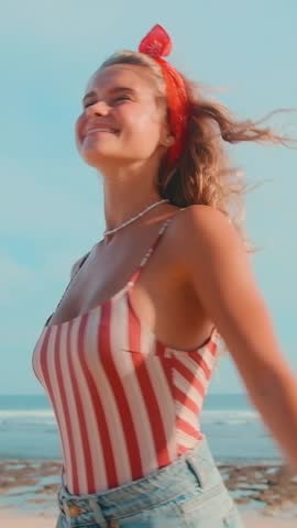 Young cheerful cute Caucasian woman teenager in denim shorts spinning stands on beach and enjoying sunshine and opportunity to indulge in getting beautiful skin tone located in tropical resort.