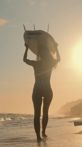Young slender woman surfer walks along sand washed by waves and holds surfboard above head after sup surfing training or competition among professional athletes located on beach at sunset.