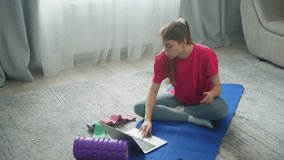 Sitting on a rug, a woman is looking for sports on a laptop. She finds the exact video and starts doing the exercises. High quality 4k footage