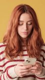 Vertical video, Red-haired woman dressed in sweater, texting on mobile phone on yellow background