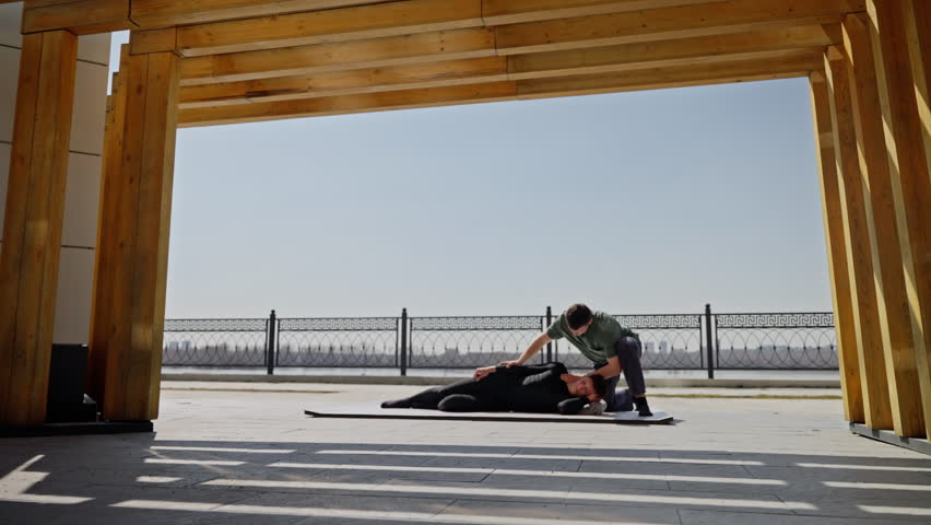 Man in green shirt performing physical therapy on person lying on mat outdoors under wooden beams with river in background.